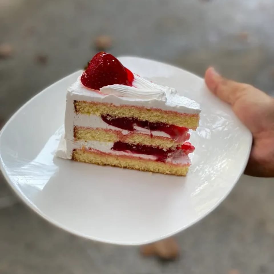 A hand holds a white plate with a slice of strawberry shortcake topped with whipped cream.