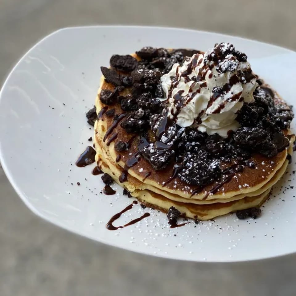 Stack of pancakes topped with whipped cream, chocolate syrup, and crushed cookies on a white plate.