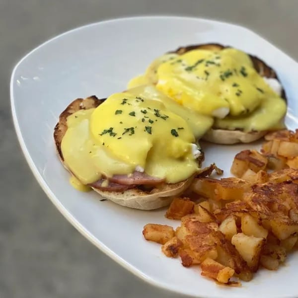 Two eggs Benedict with hollandaise sauce and a side of crispy home fries on a white plate.