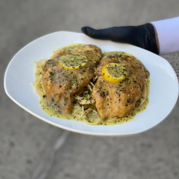 Two lemon chicken breasts with herbs and sauce over spaghetti, held by a gloved hand.