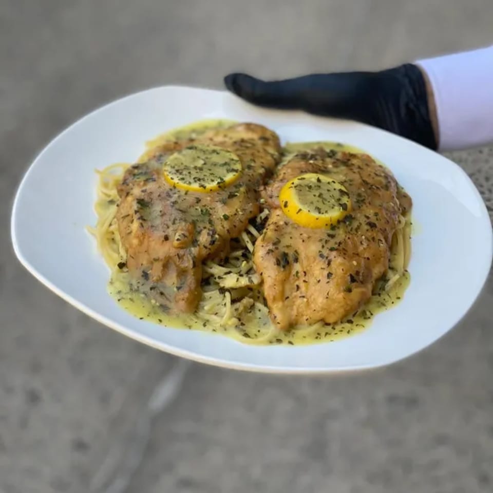 Two lemon chicken breasts with herbs and sauce over spaghetti, held by a gloved hand.