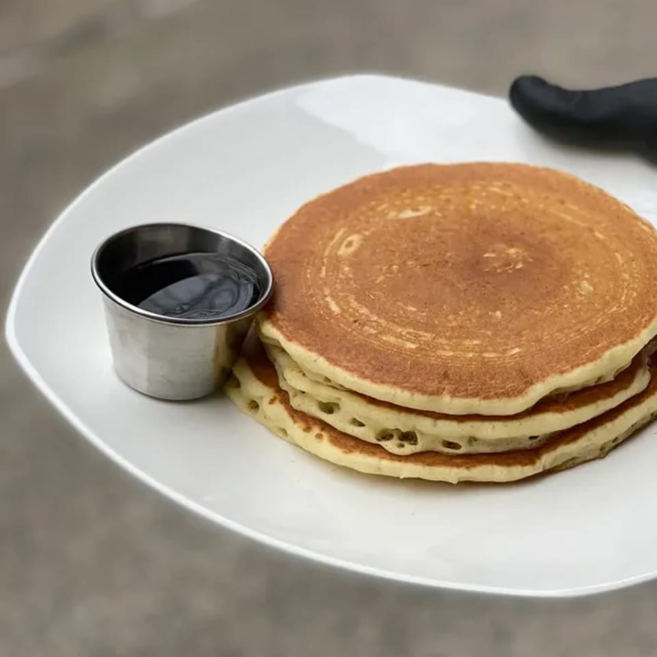 A stack of three golden pancakes with a side of syrup in a metal cup on a white plate.