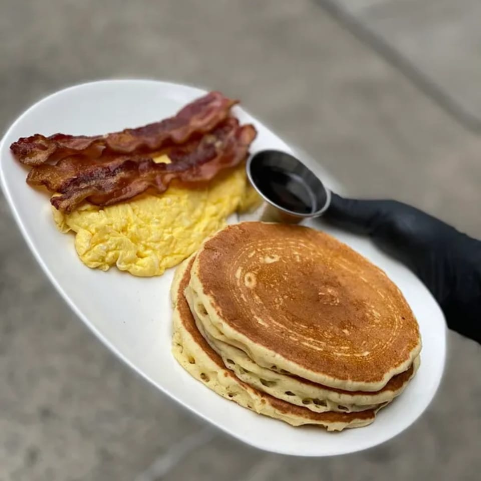 A Squire's Diner breakfast plate with scrambled eggs, crispy bacon, pancakes, and a side of syrup.