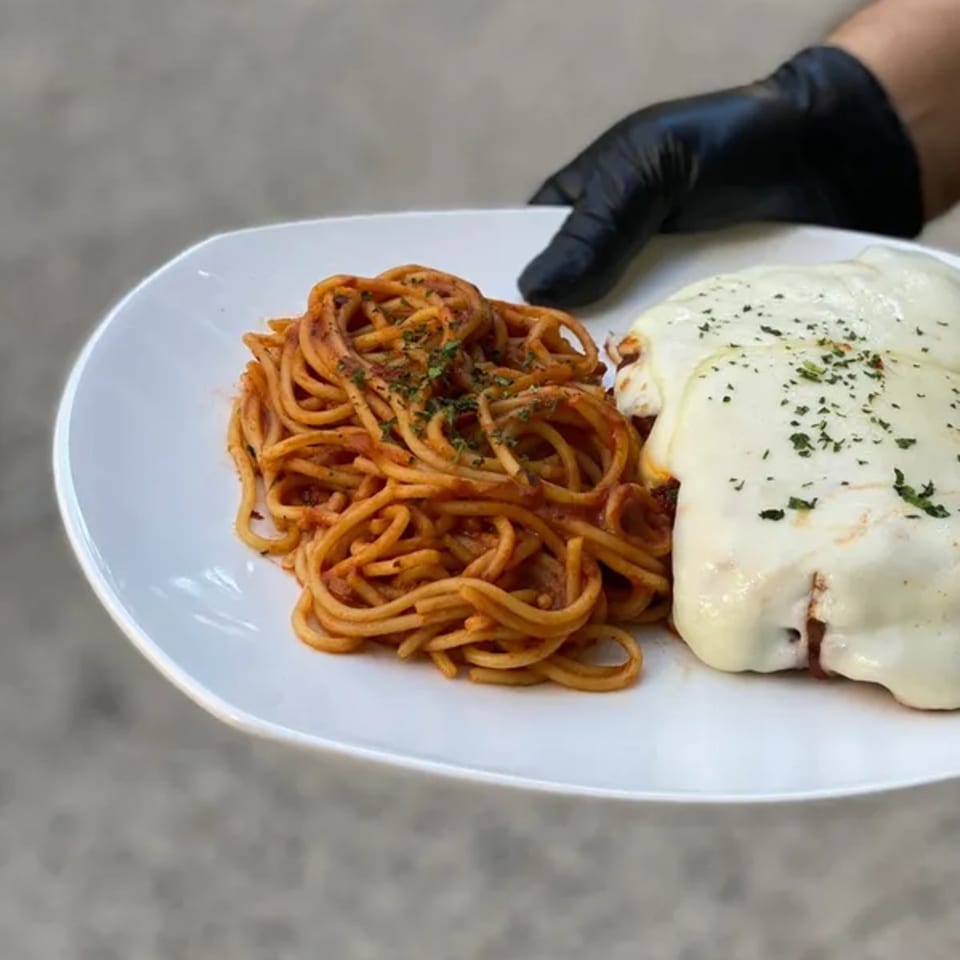 A hand in a black glove holds a white plate with spaghetti and chicken parmesan.